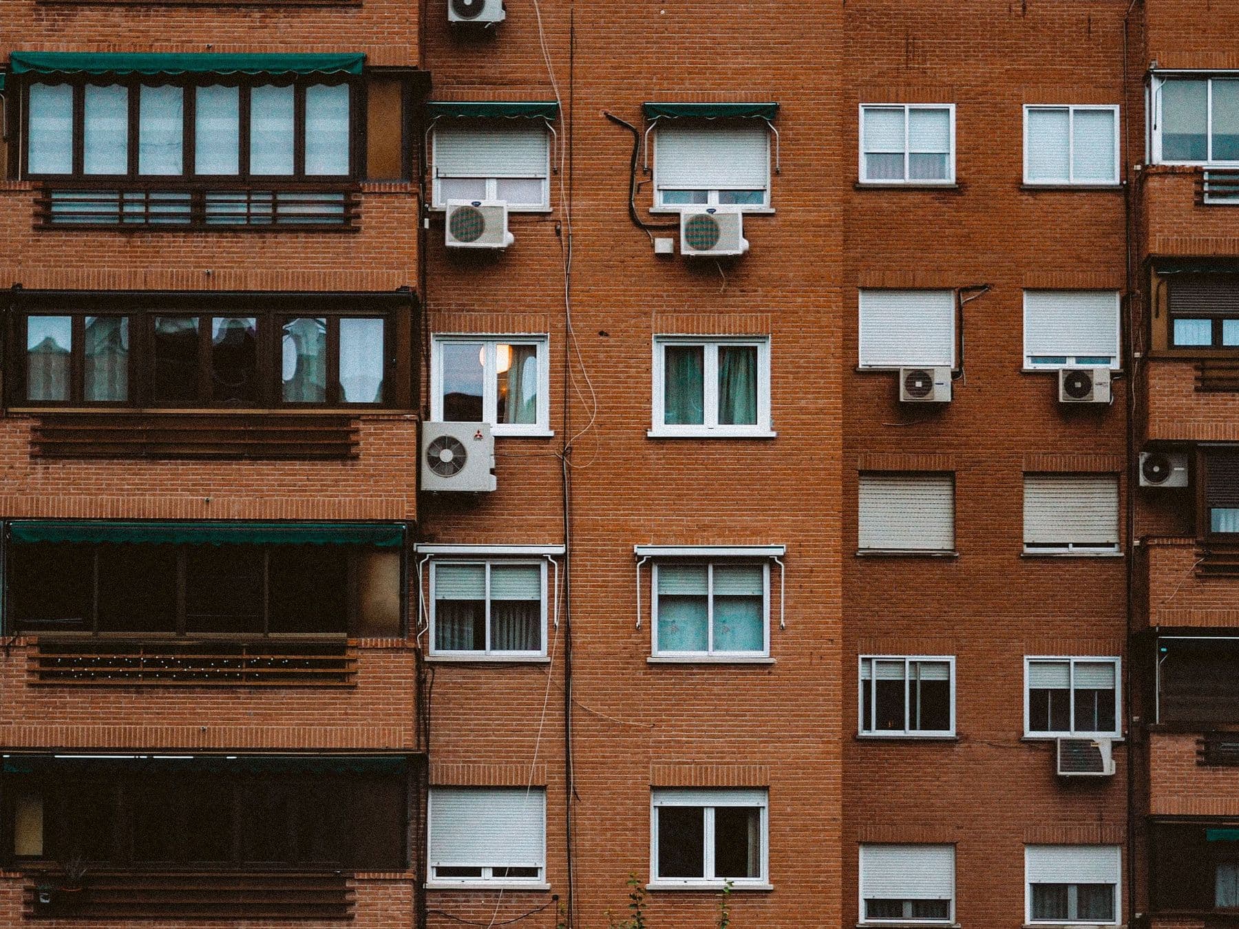 Fachada de ladrillo de un bloque de pisos con persianas enrollables a distintas alturas, terrazas cerradas y aparatos de aire acondicionado