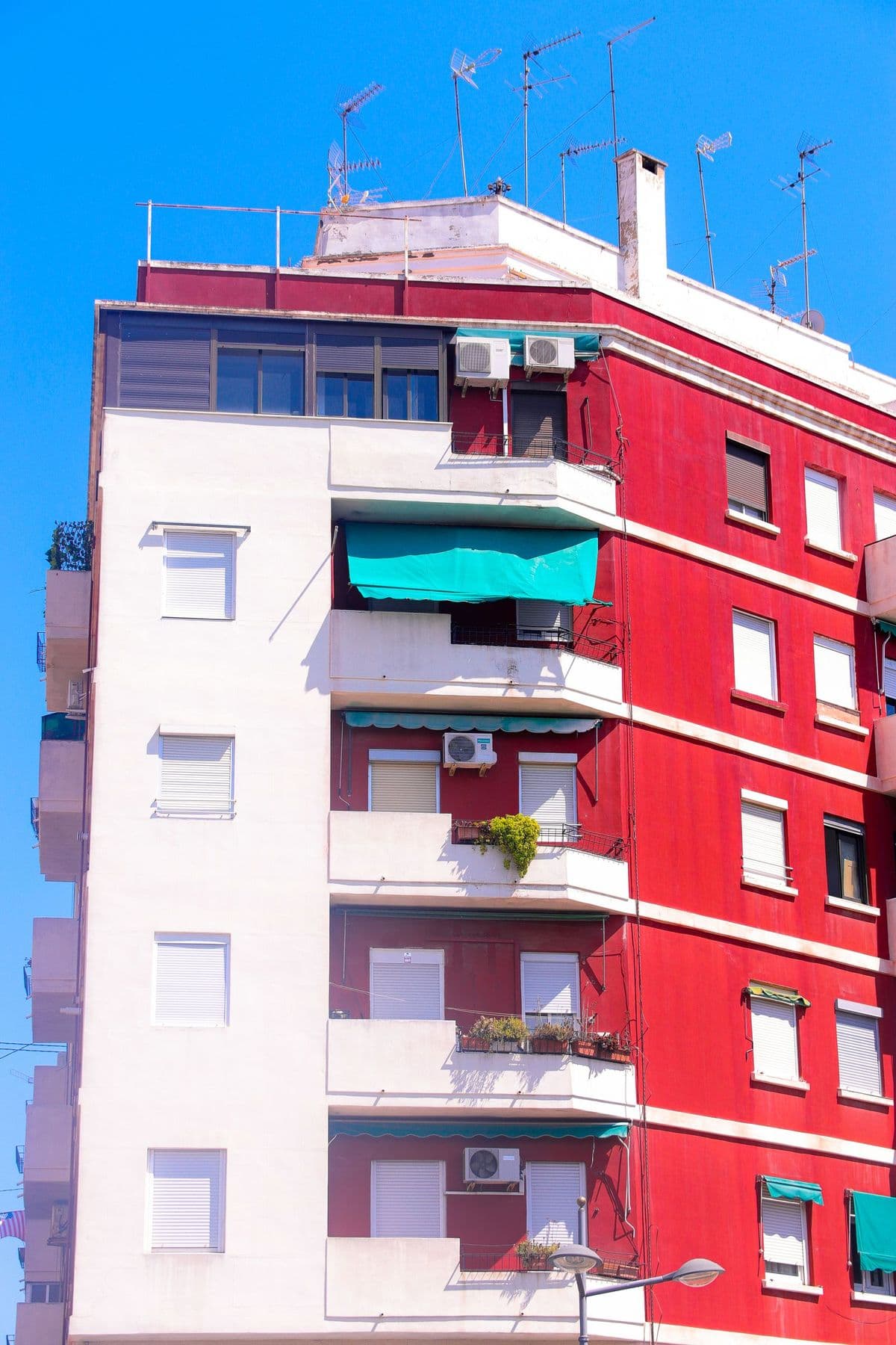 Edificio de viviendas rojo y blanco con persianas enrollables, un toldo turquesa en un balcón, antenas en la azotea y cielo azul intenso