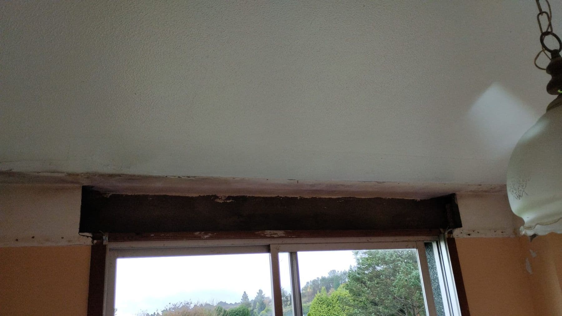 Cajón de persiana desmontado encima de una ventana corredera, con el hueco de obra a la vista y árboles al otro lado del cristal
