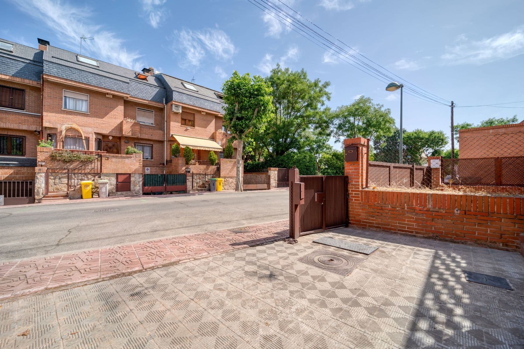 Calle de chalets adosados de ladrillo con toldos, tejados de pizarra y árboles, vista desde una entrada con verja bajo un cielo de verano