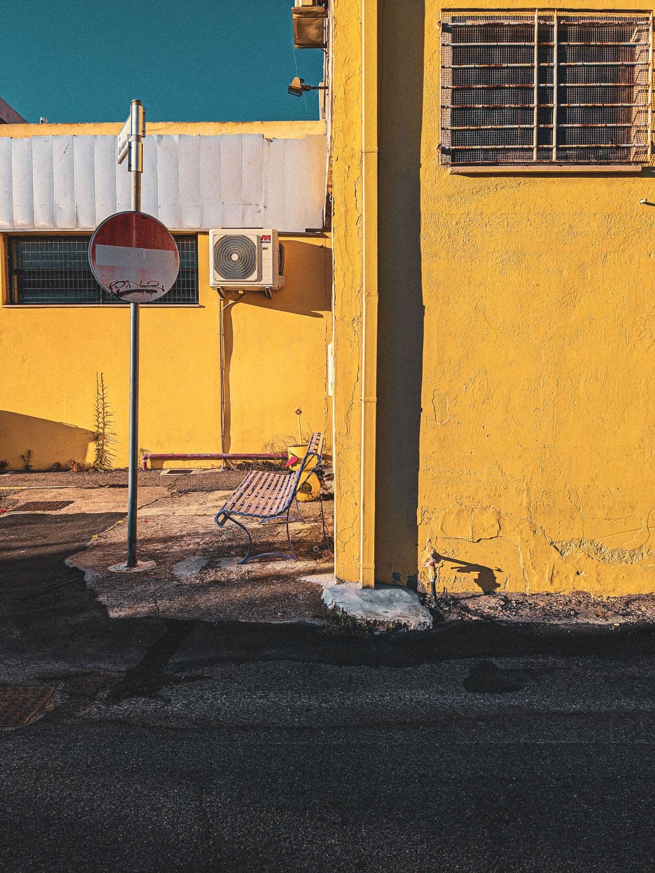 Calle con sol intenso y sombras duras sobre una pared amarilla, con una señal de dirección prohibida, un banco y un aire acondicionado