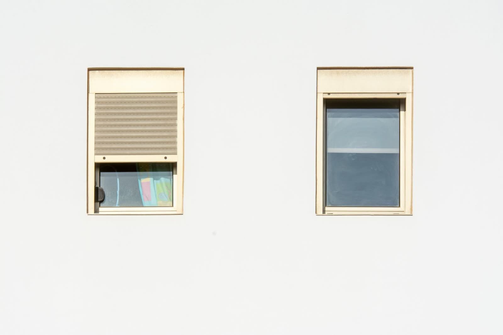 Fachada blanca lisa con dos ventanas pequeñas, una con la persiana bajada hasta dos tercios y la otra con la persiana recogida en el cajón
