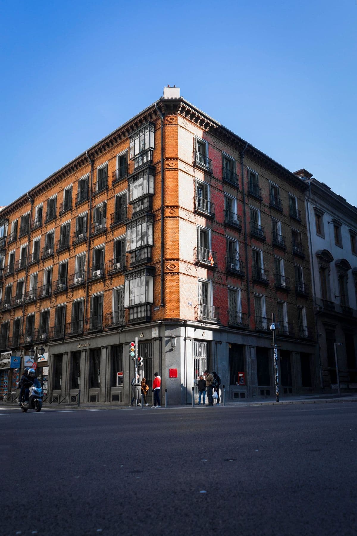 Edificio residencial de ladrillo en esquina con miradores y balcones, visto desde el asfalto de un cruce con peatones y una moto a pie de calle