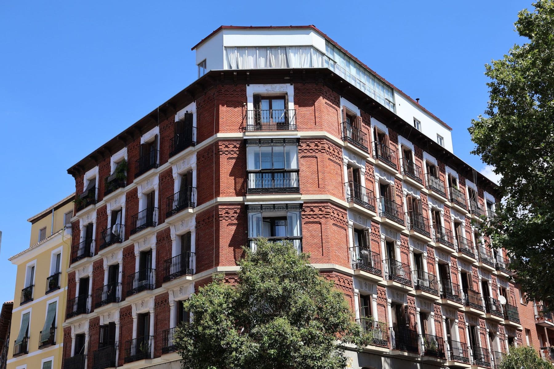 Edificio de viviendas de ladrillo rojo en esquina, con balcones de forja, miradores acristalados y árboles delante bajo un cielo azul despejado