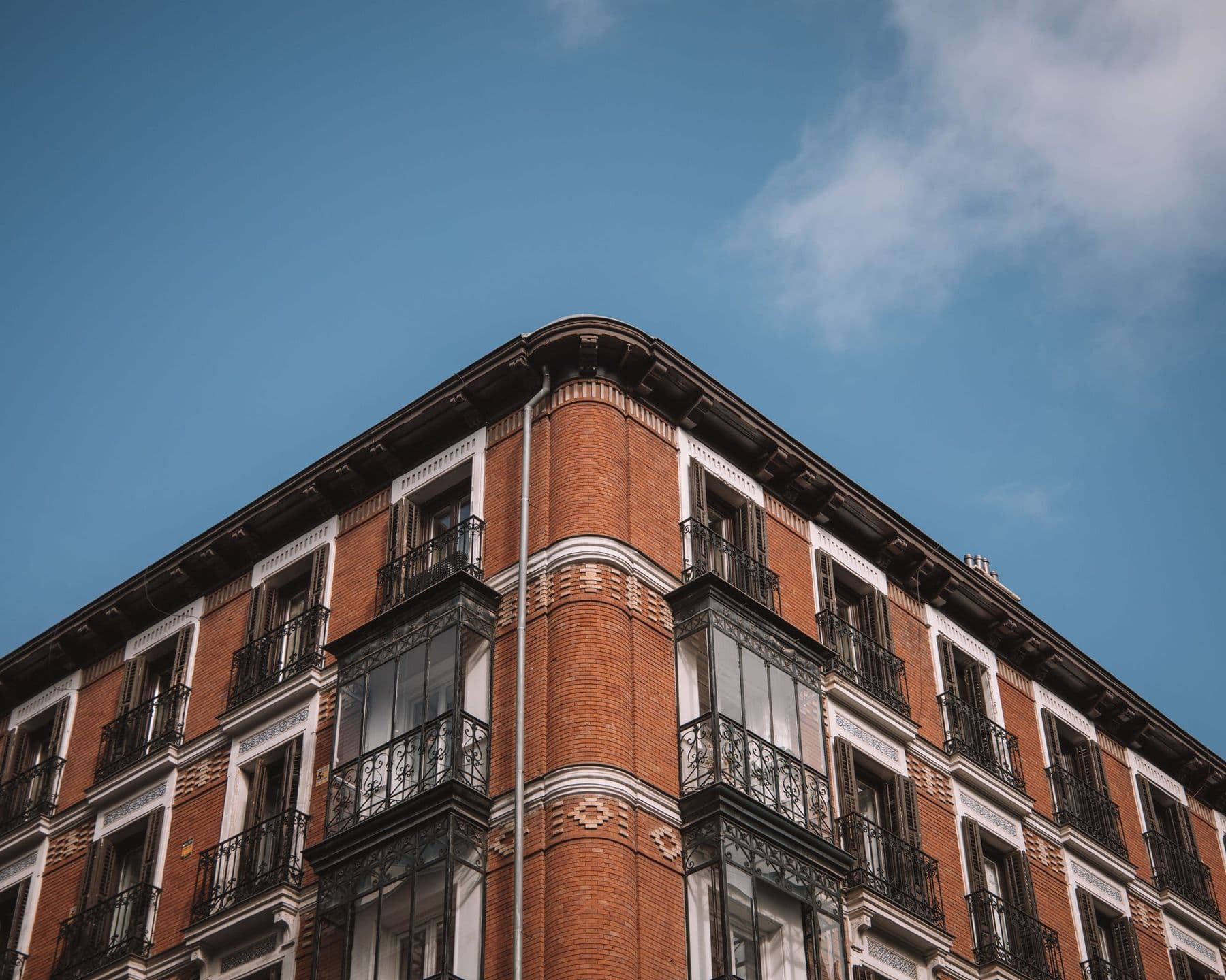 Esquina de un edificio de ladrillo con miradores de hierro, balcones, bandas decorativas blancas y cornisa oscura vista en contrapicado contra un cielo azul