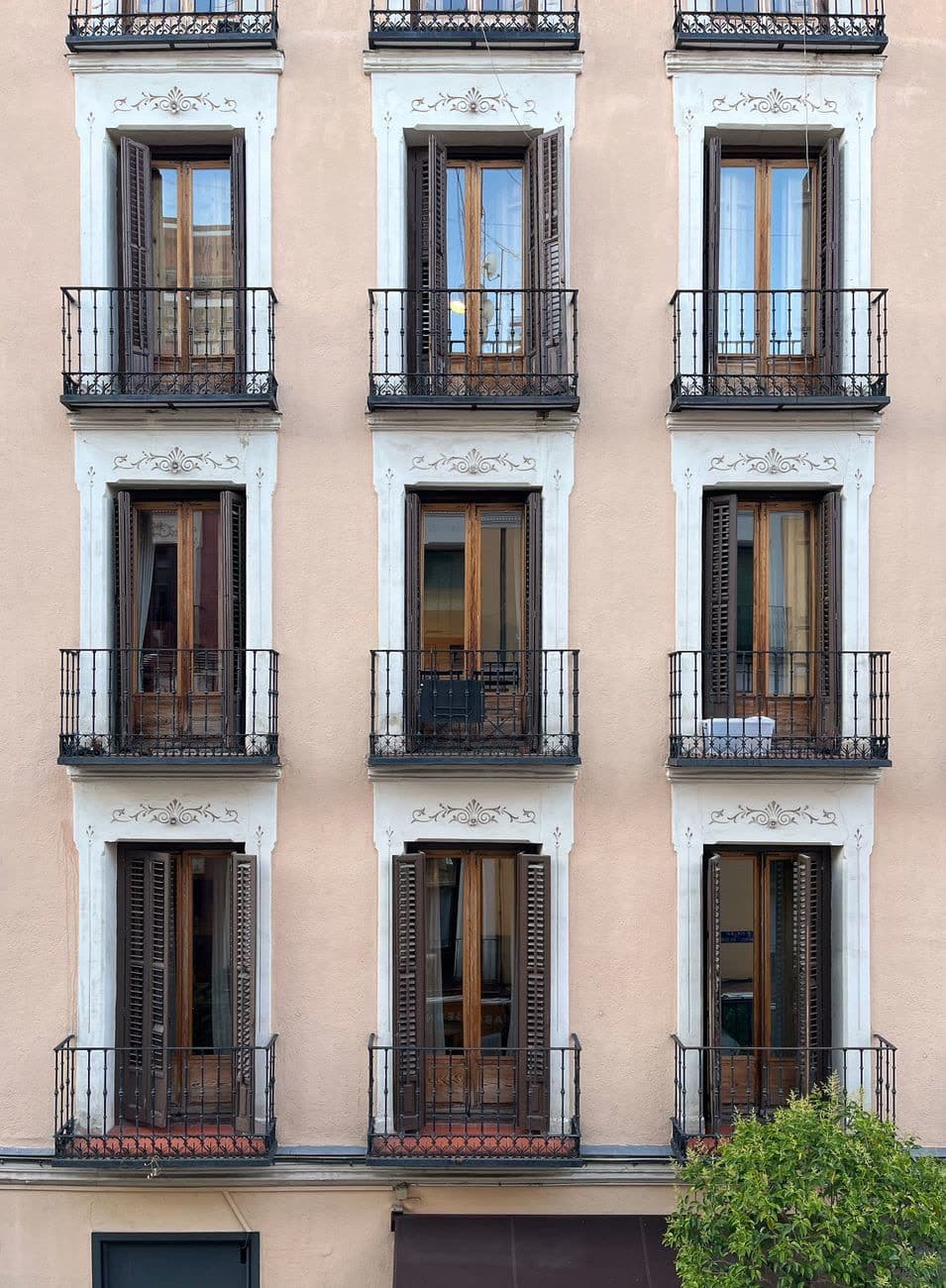 Fachada clásica rosada con tres columnas de balcones de forja, molduras blancas y contraventanas de madera abiertas o entornadas