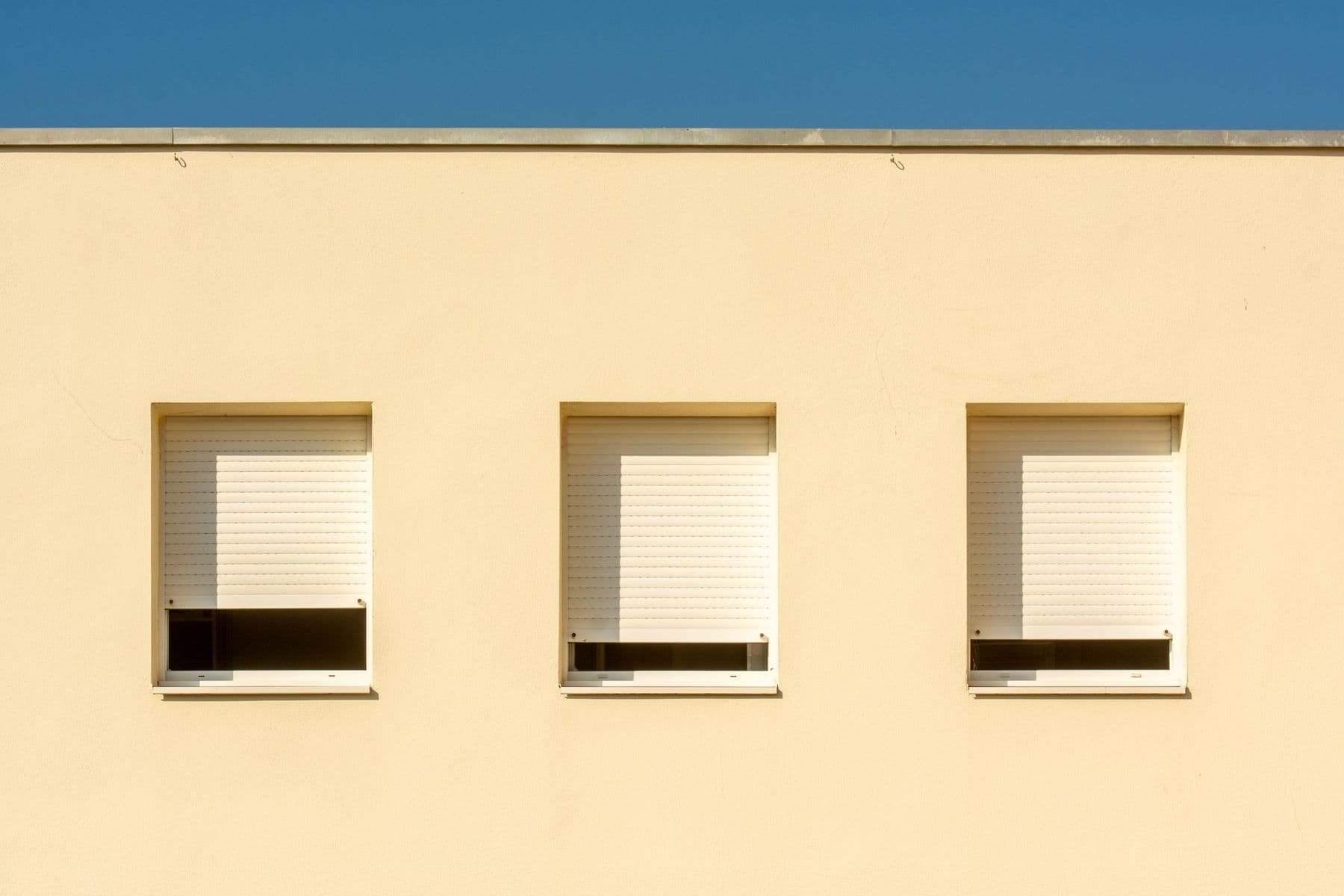 Fachada lisa de color crema con tres ventanas iguales y sus persianas blancas bajadas casi hasta abajo bajo una franja de cielo azul