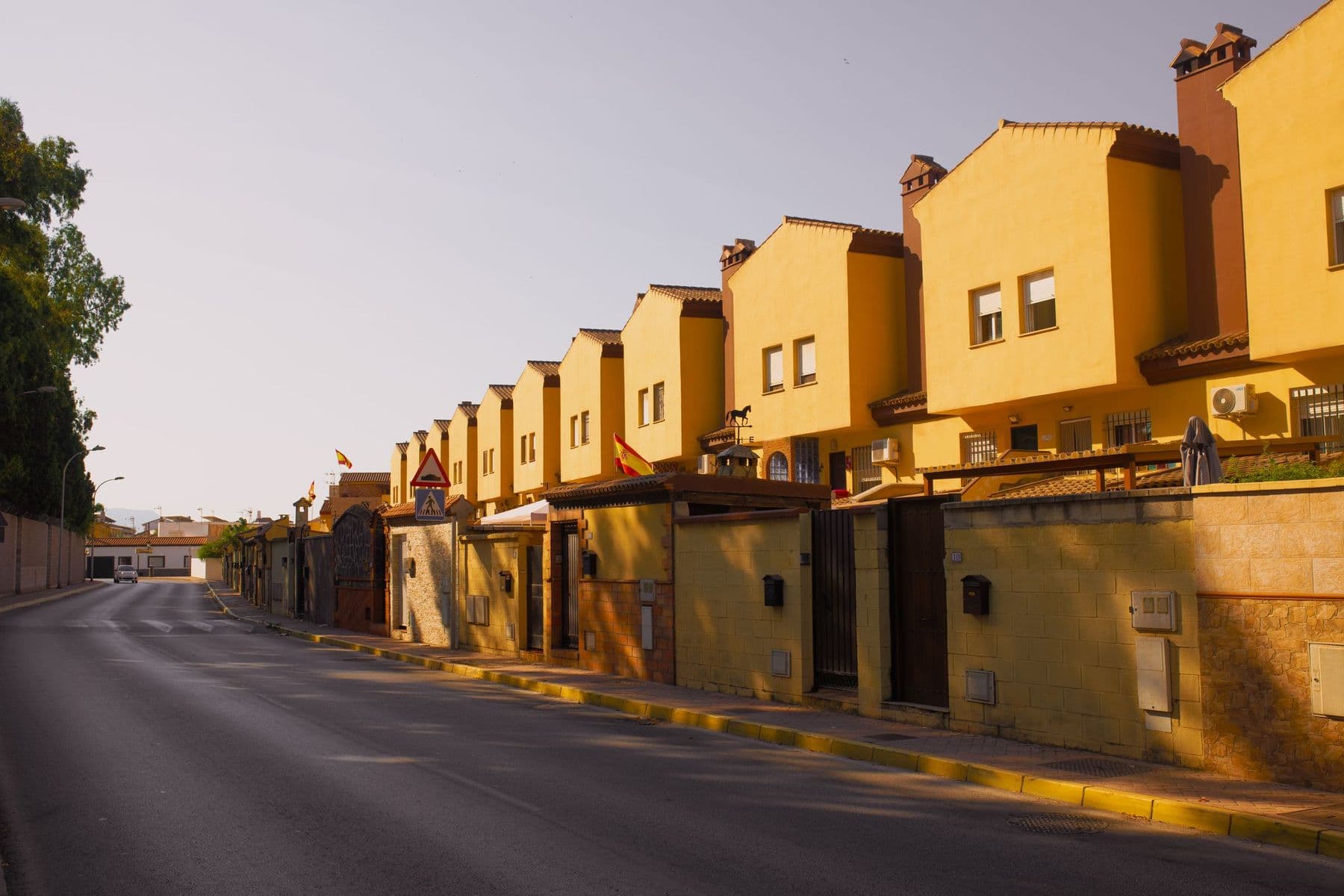 Hilera de chalets adosados de fachada amarilla con chimeneas y muros de entrada a lo largo de una calle vacía con luz de atardecer