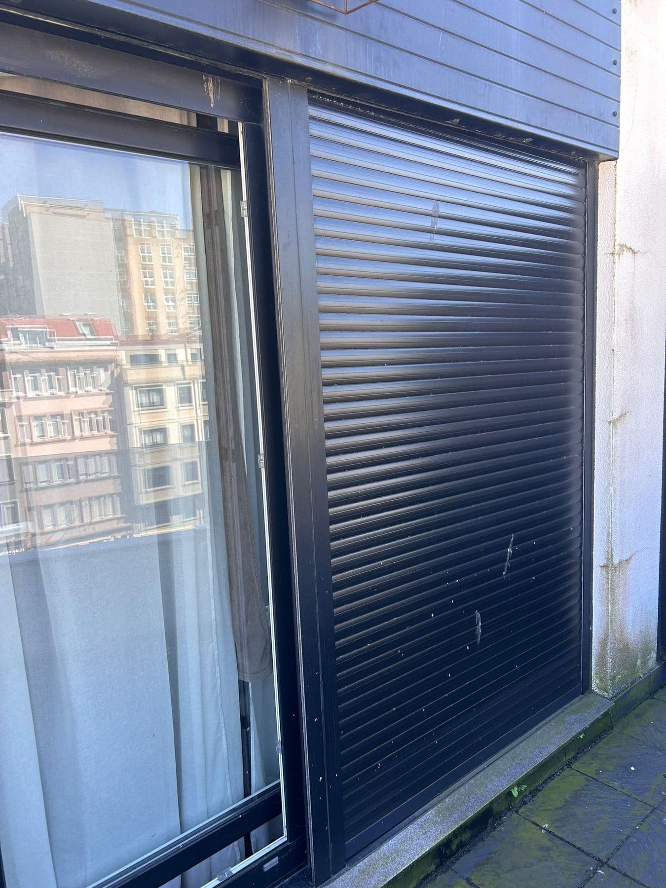 Persiana enrollable de aluminio oscuro bajada del todo en una balconera, junto a la hoja corredera de cristal y el suelo de la terraza con musgo