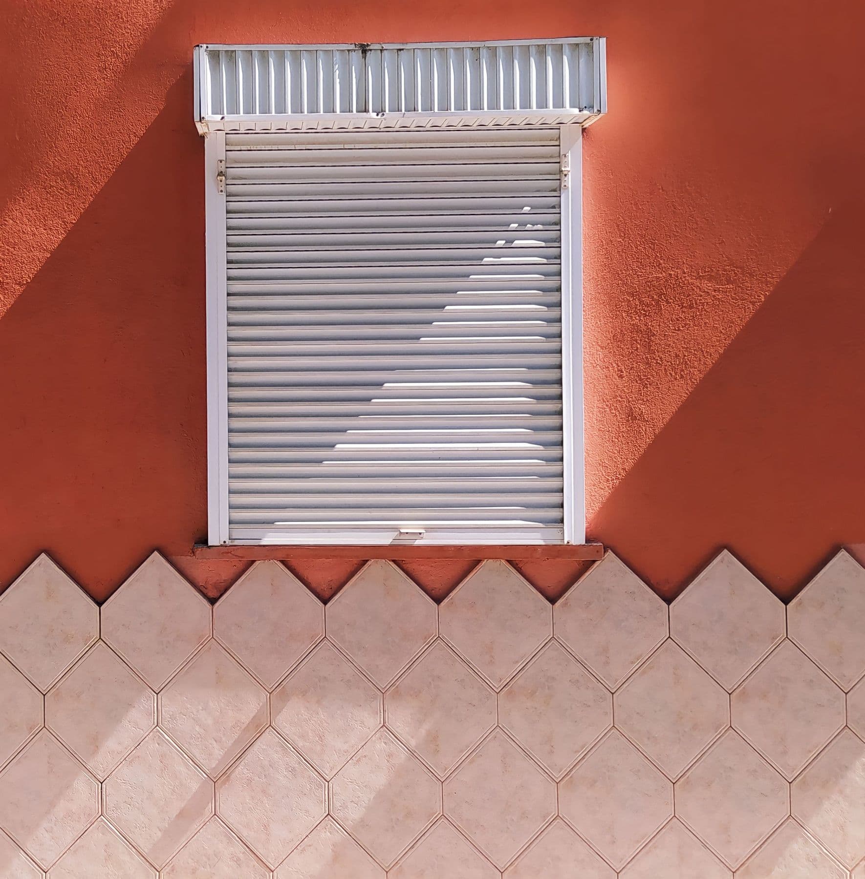 Persiana enrollable blanca bajada en una ventana de pared roja, con una sombra diagonal sobre las lamas y un zócalo de azulejos debajo