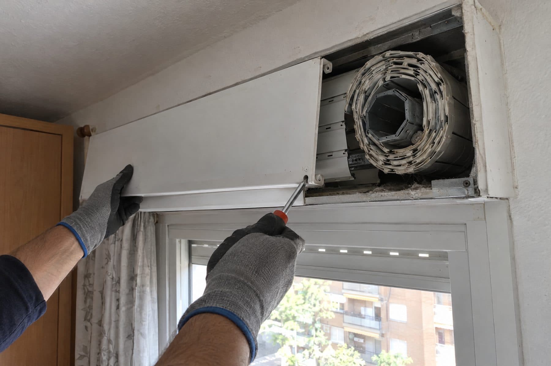 Manos con guantes de trabajo retirando con un destornillador la tapa interior del cajón de una persiana, que deja ver el eje octogonal y el paño enrollado