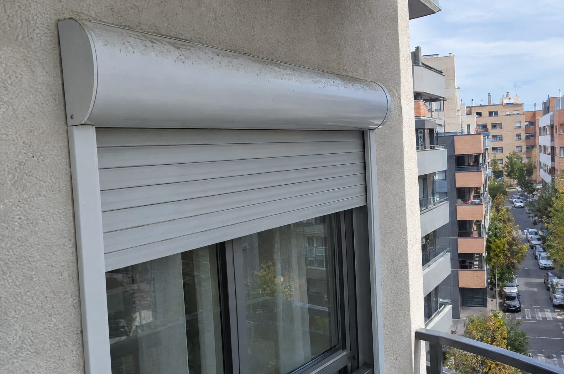 Cajón compacto de aluminio montado por fuera sobre la ventana de un piso moderno, con guías del mismo color y la persiana a media altura