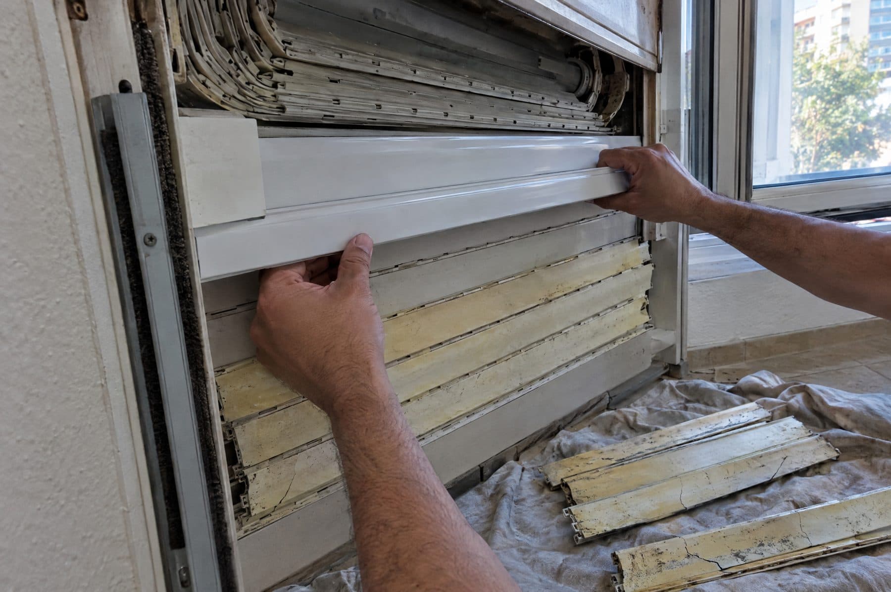 Manos de un técnico metiendo de lado una lama nueva de PVC en el paño de una persiana, con lamas viejas amarillentas y rajadas sobre una lona en el suelo
