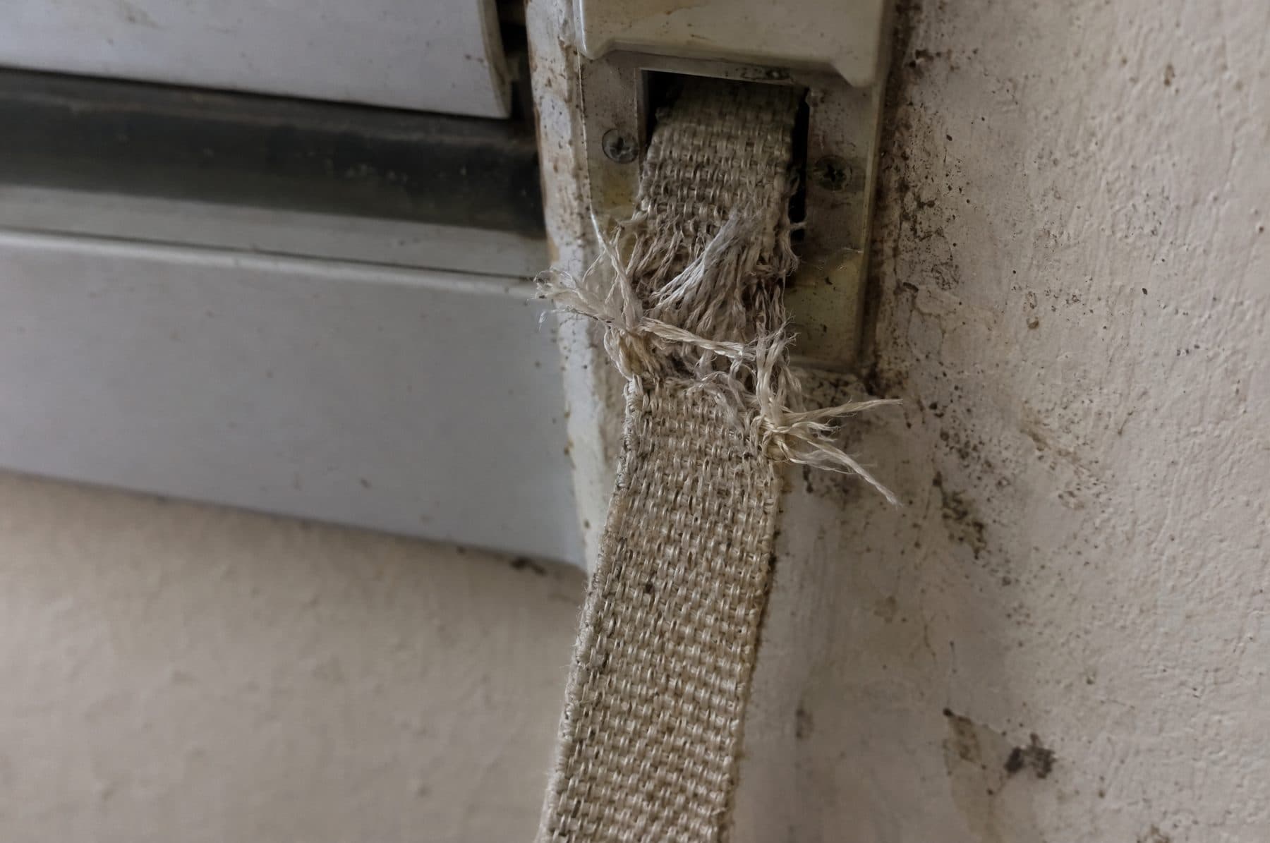 Cinta de persiana beige muy deshilachada justo donde entra en la guía de entrada de la pared, bajo la ventana