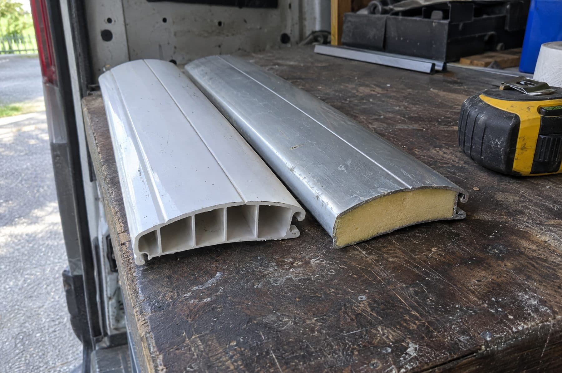 Dos lamas de persiana sobre un banco de trabajo: una de PVC blanco con las cámaras huecas a la vista en el corte y otra de aluminio con el relleno de espuma de poliuretano