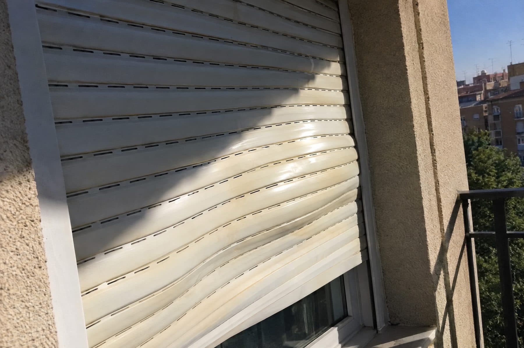 Persiana de PVC blanco bajada con las lamas onduladas y abombadas por el calor, al sol de la tarde en un balcón