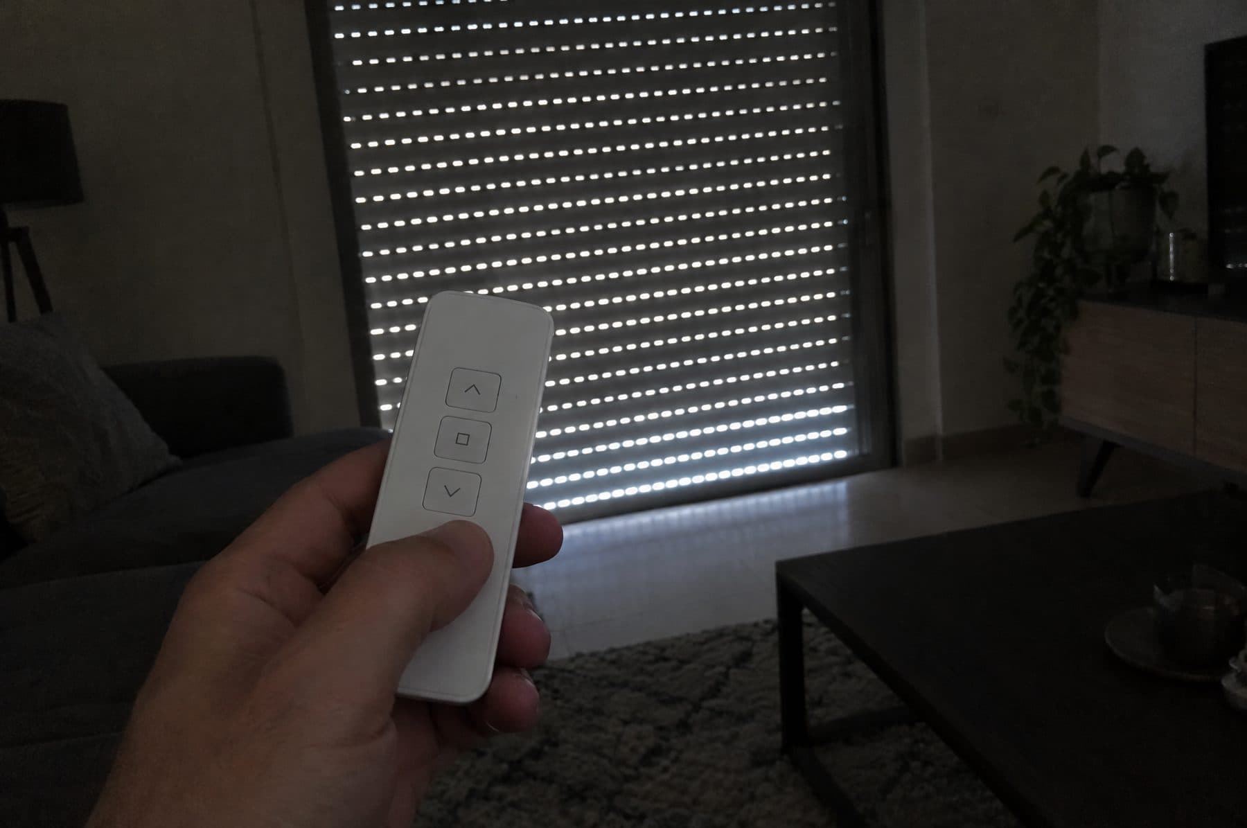 Mano sujetando un mando blanco de persiana motorizada en un salón en penumbra, con la persiana bajada y rendijas de luz entre las lamas