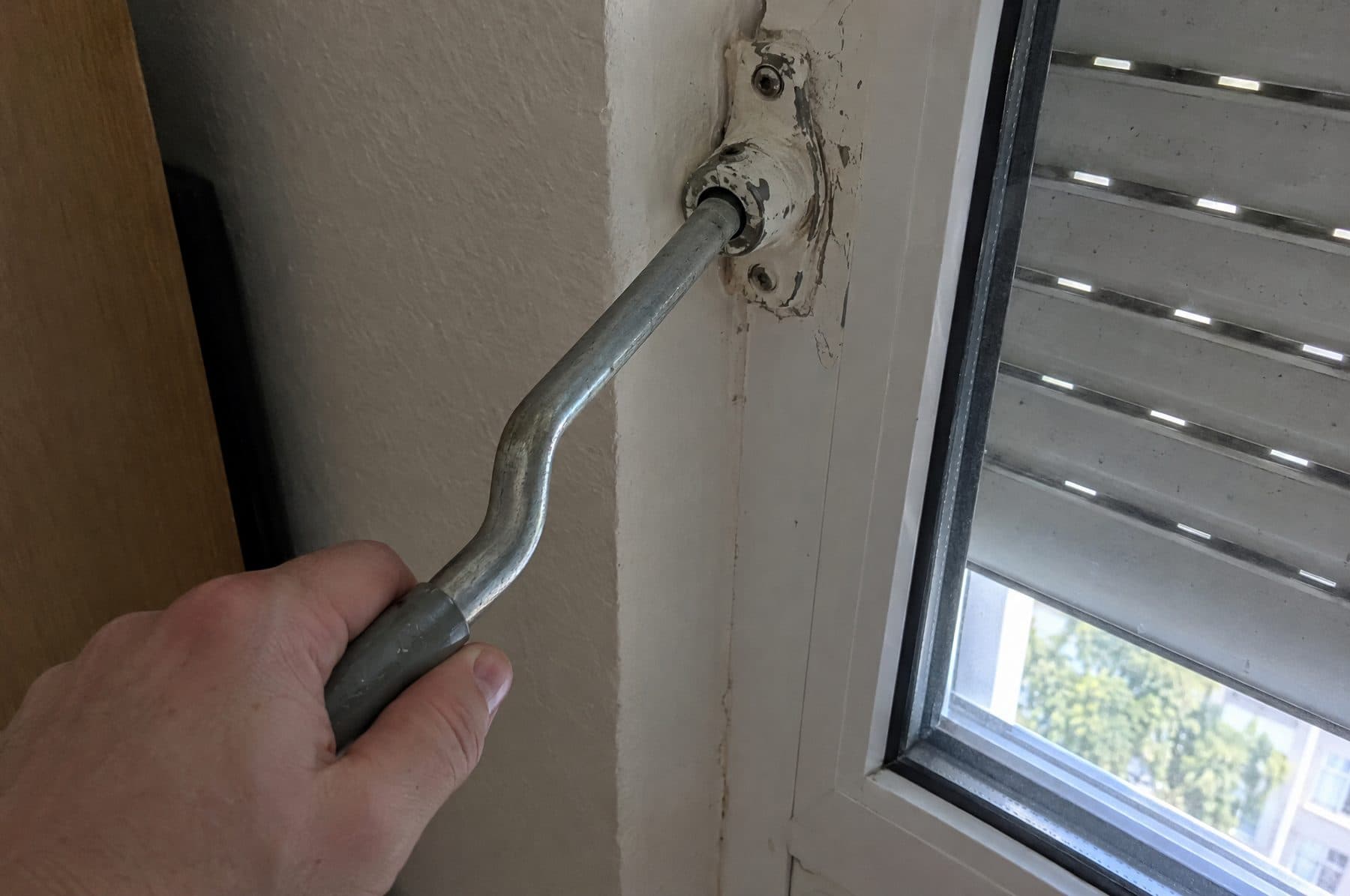 Mano girando la manivela de una persiana enganchada a la rótula que sale de la pared junto a la ventana