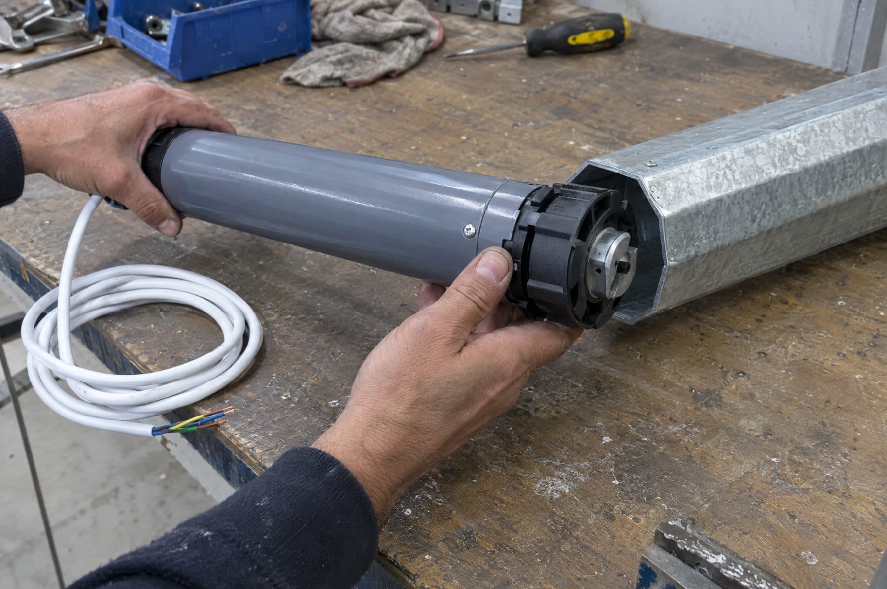 Manos de un técnico introduciendo un motor tubular gris en un eje octogonal de acero galvanizado sobre un banco de trabajo, con el cable blanco enrollado