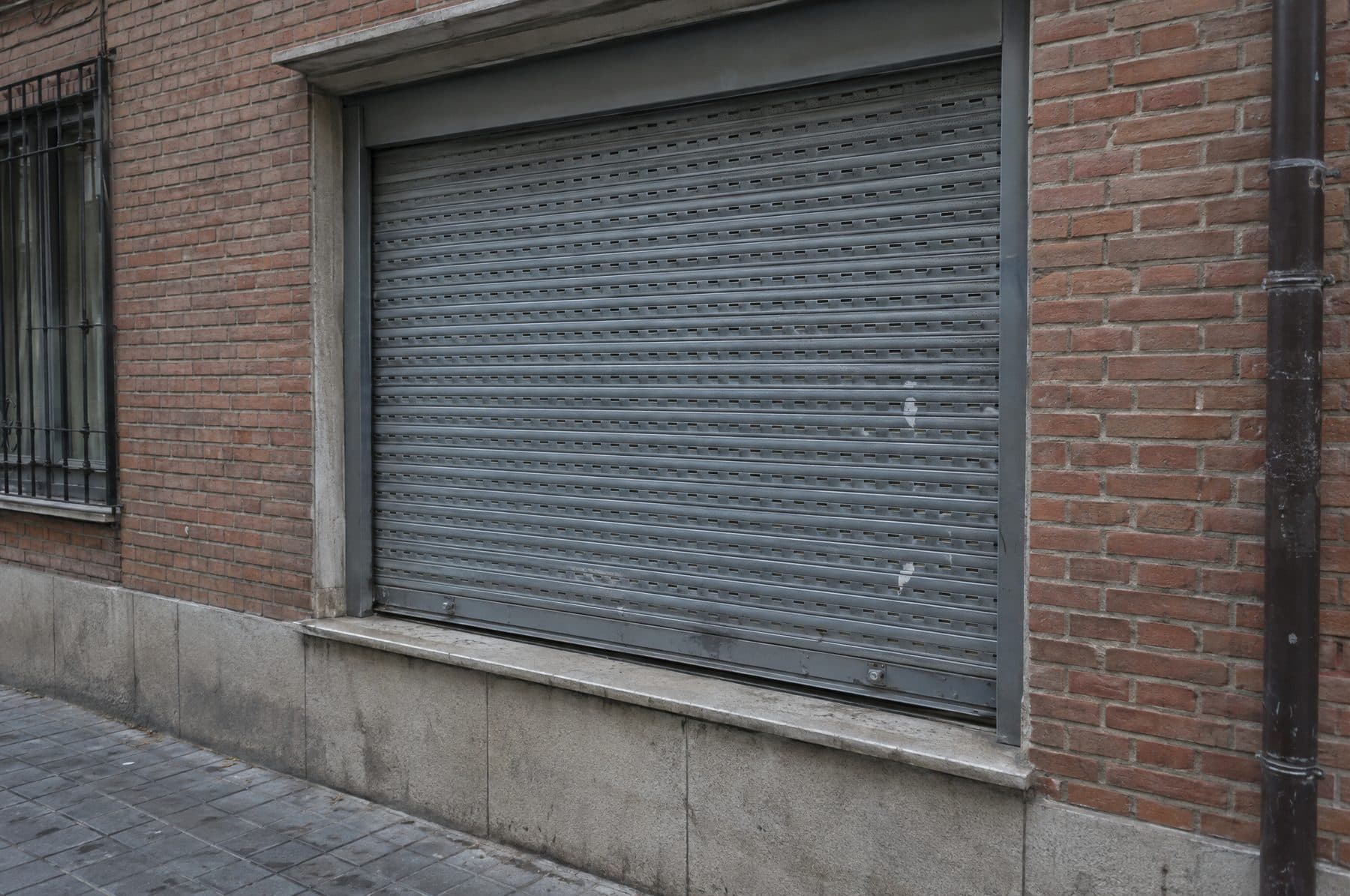 Ventana de planta baja en un edificio de ladrillo cerrada con una persiana gris de aluminio extrusionado, con pequeñas abolladuras y suciedad de calle