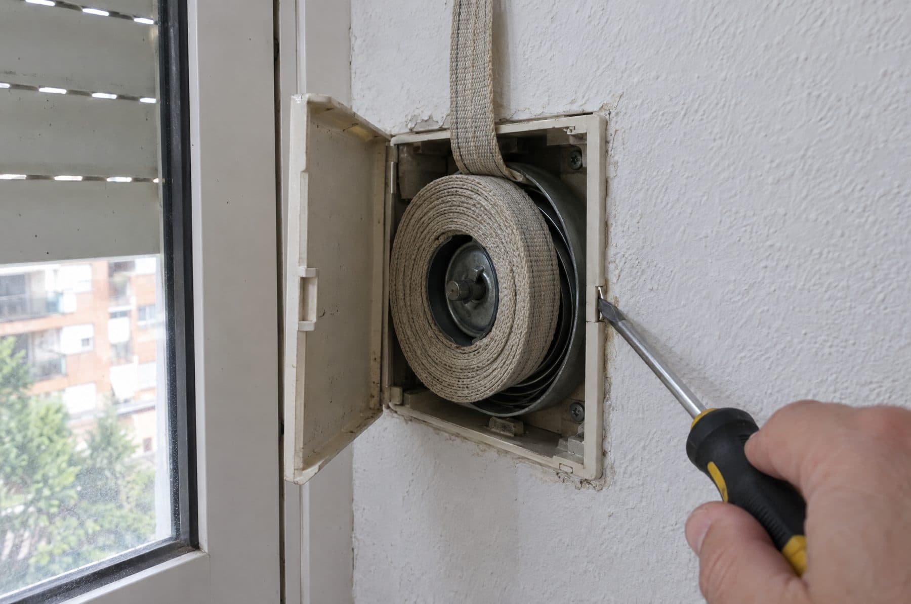 Recogedor de cinta empotrado en la pared junto a una ventana, con la tapa abierta, la cinta enrollada en el tambor del muelle y un destornillador apoyado en el borde